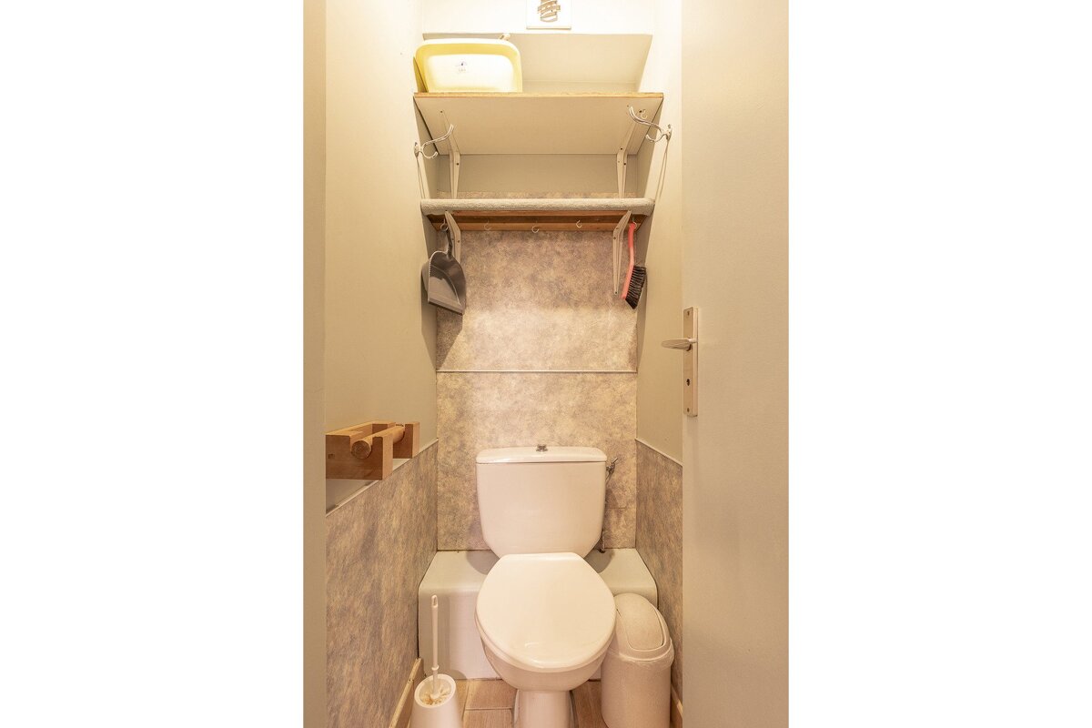A compact toilet room featuring a white toilet, tiled walls, shelves with cleaning tools, a toilet brush, and a small trash can.