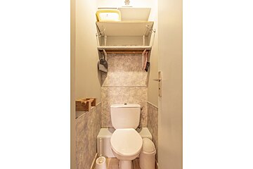 A compact toilet room featuring a white toilet, tiled walls, shelves with cleaning tools, a toilet brush, and a small trash can.