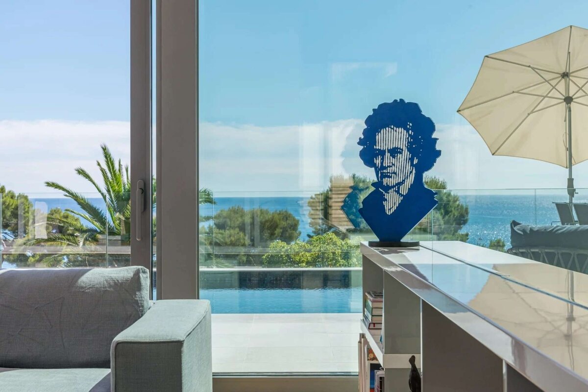 A blue Beethoven bust sits in a modern room with large windows, overlooking an infinity pool and the sparkling ocean, with palm trees and a patio umbrella.