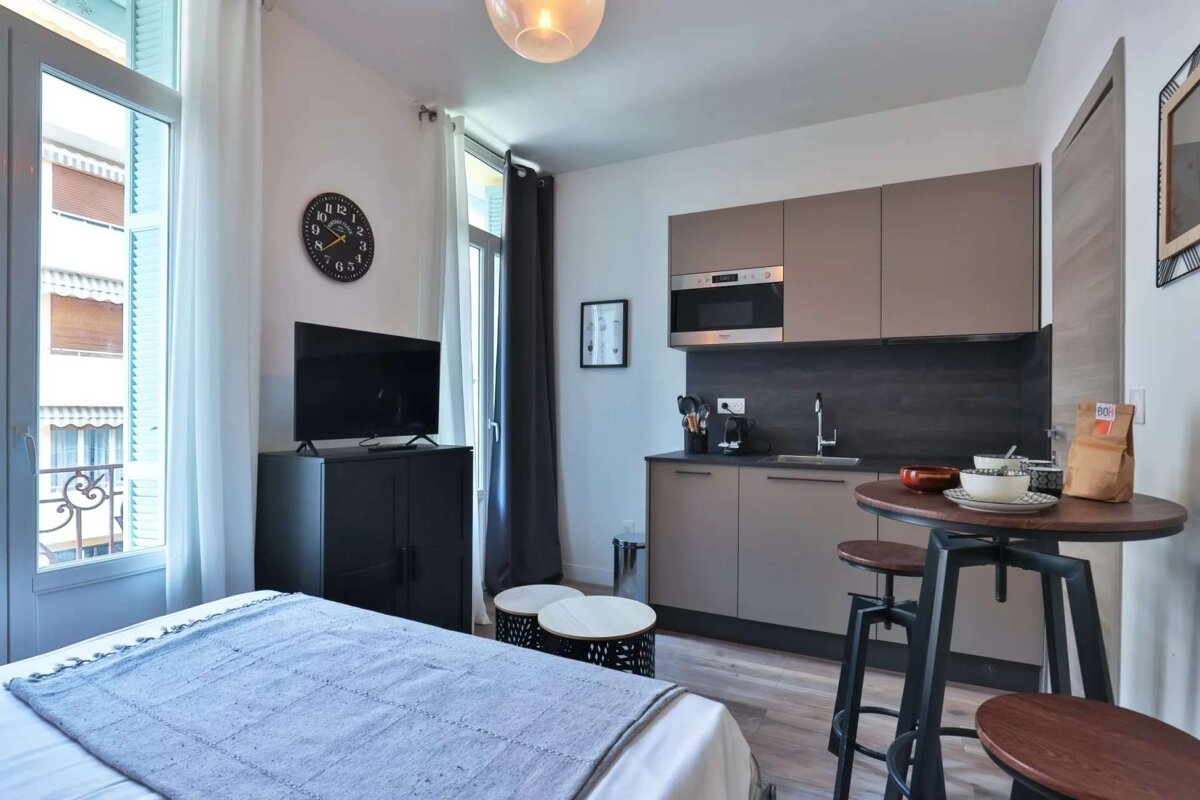 A modern studio apartment features a bed, a compact kitchenette, and a high-top dining table. A TV and balcony door are also visible.