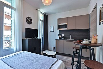 A modern studio apartment features a bed, a compact kitchenette, and a high-top dining table. A TV and balcony door are also visible.