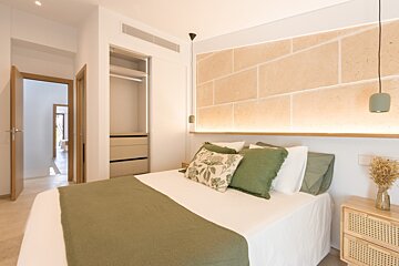 Cozy bedroom with a white bed, green textiles, a stone headboard with ambient lighting, an open closet, and a rattan nightstand.