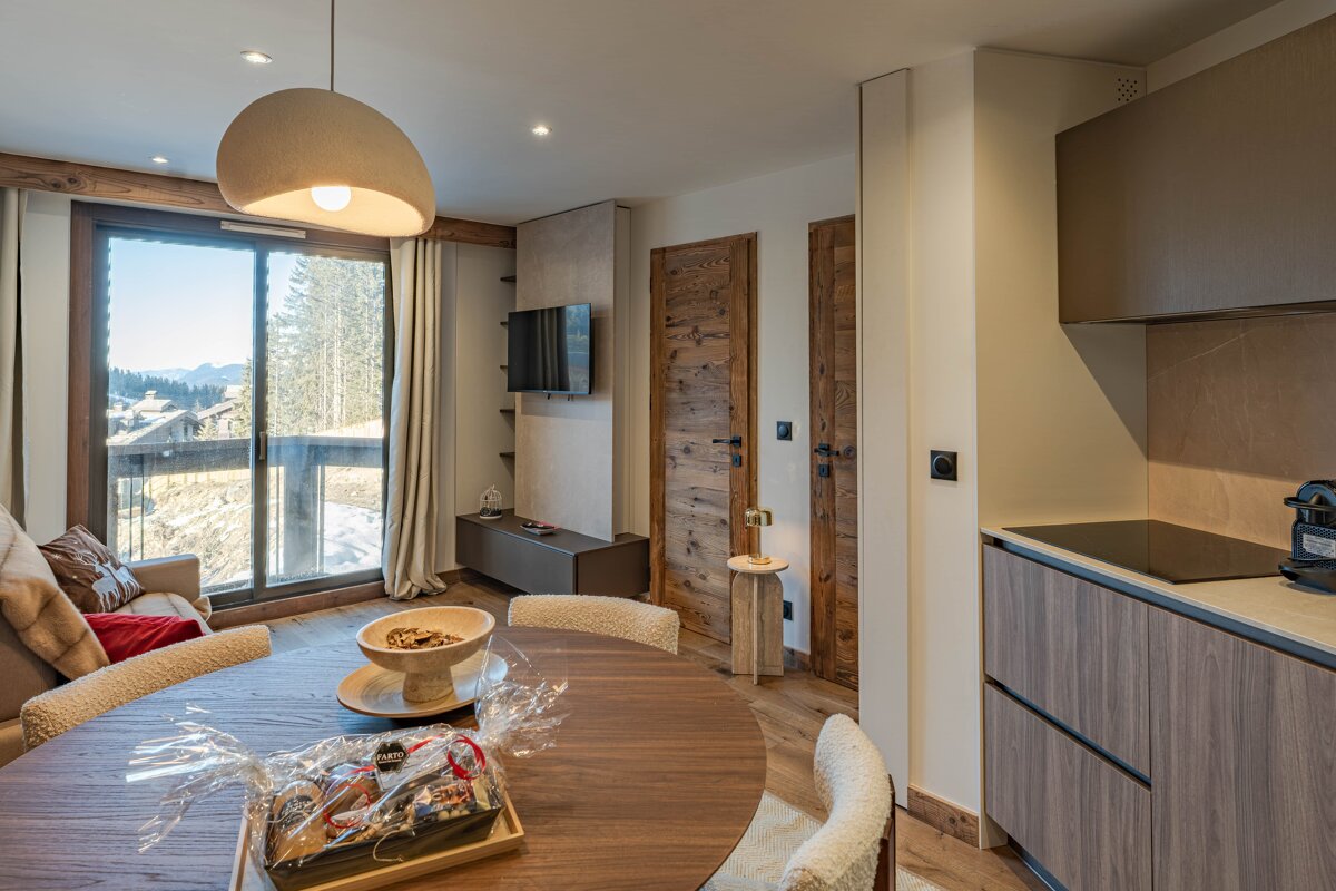 A modern, cozy living space with a round dining table, sofa, kitchenette, and a large window overlooking snowy mountains.
