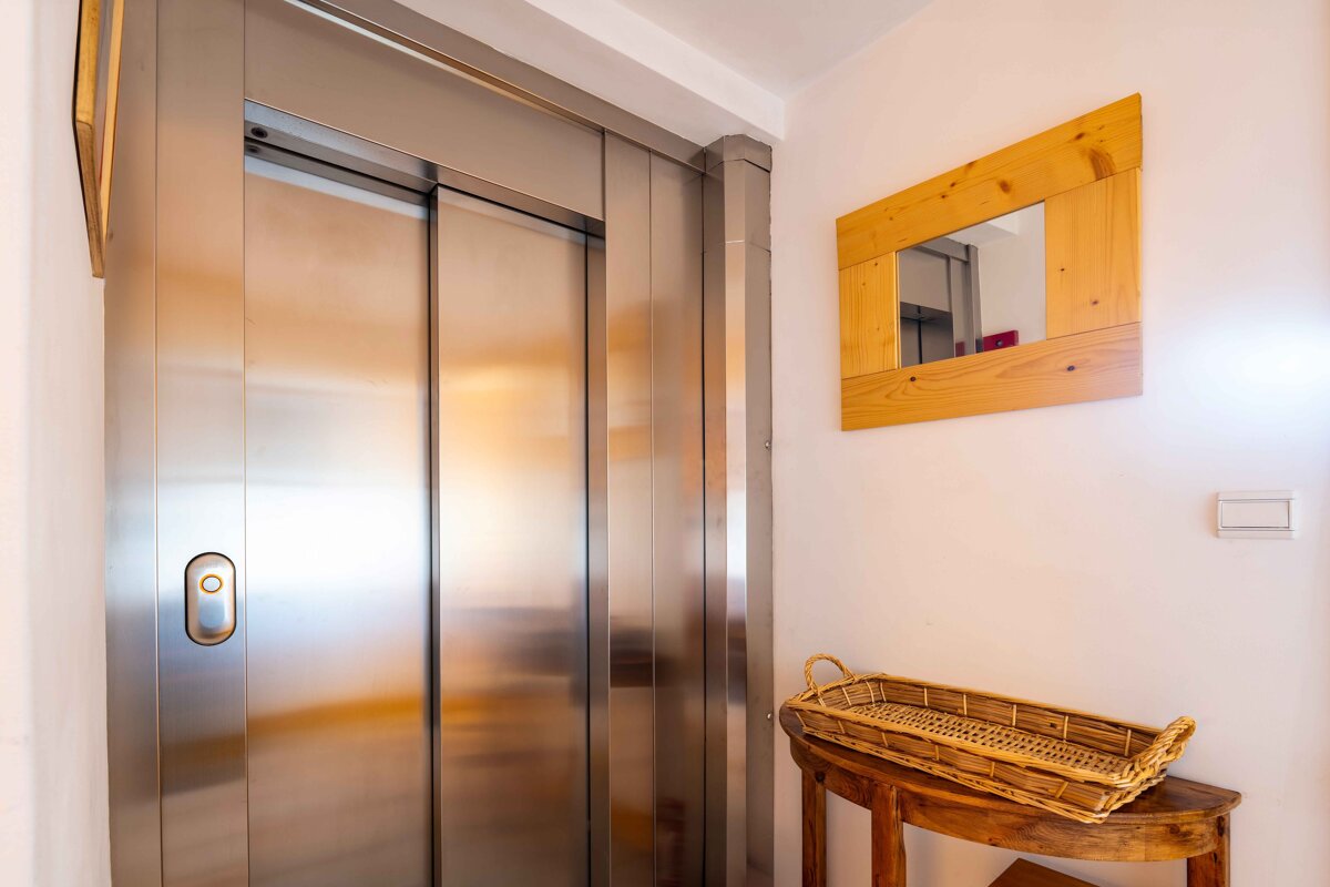 Stainless steel elevator doors with a call button, beside a wooden mirror and a wooden table with a woven basket.
