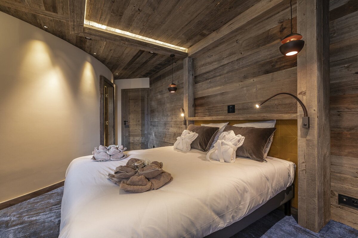 Cozy bedroom with rustic wood walls/ceiling, a bed draped in white linen and adorned with folded robes/towels. Warm ambient and task lighting completes the look.