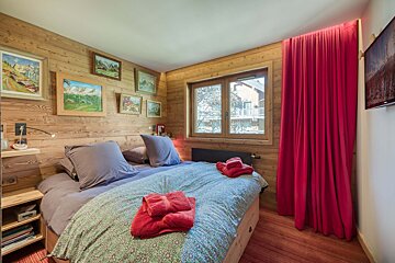 A warm, wood-paneled bedroom with a patterned bed, red towels, and matching curtains. Mountain paintings decorate the walls, and a window shows the outside.