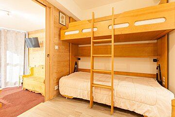 A cozy room with light wooden bunk beds; the bottom bed pulls out. Wood-paneled walls and an adjacent room with a TV and bench are visible.