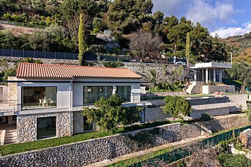Modern house with red tile roof on a terraced hillside, featuring stone walls, lush greenery, and a pool area under a blue sky.