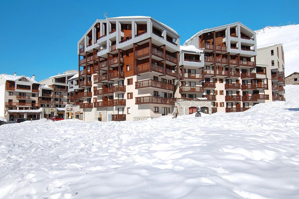 A snow-covered alpine resort with modern multi-story buildings featuring wooden balconies under a clear blue sky.