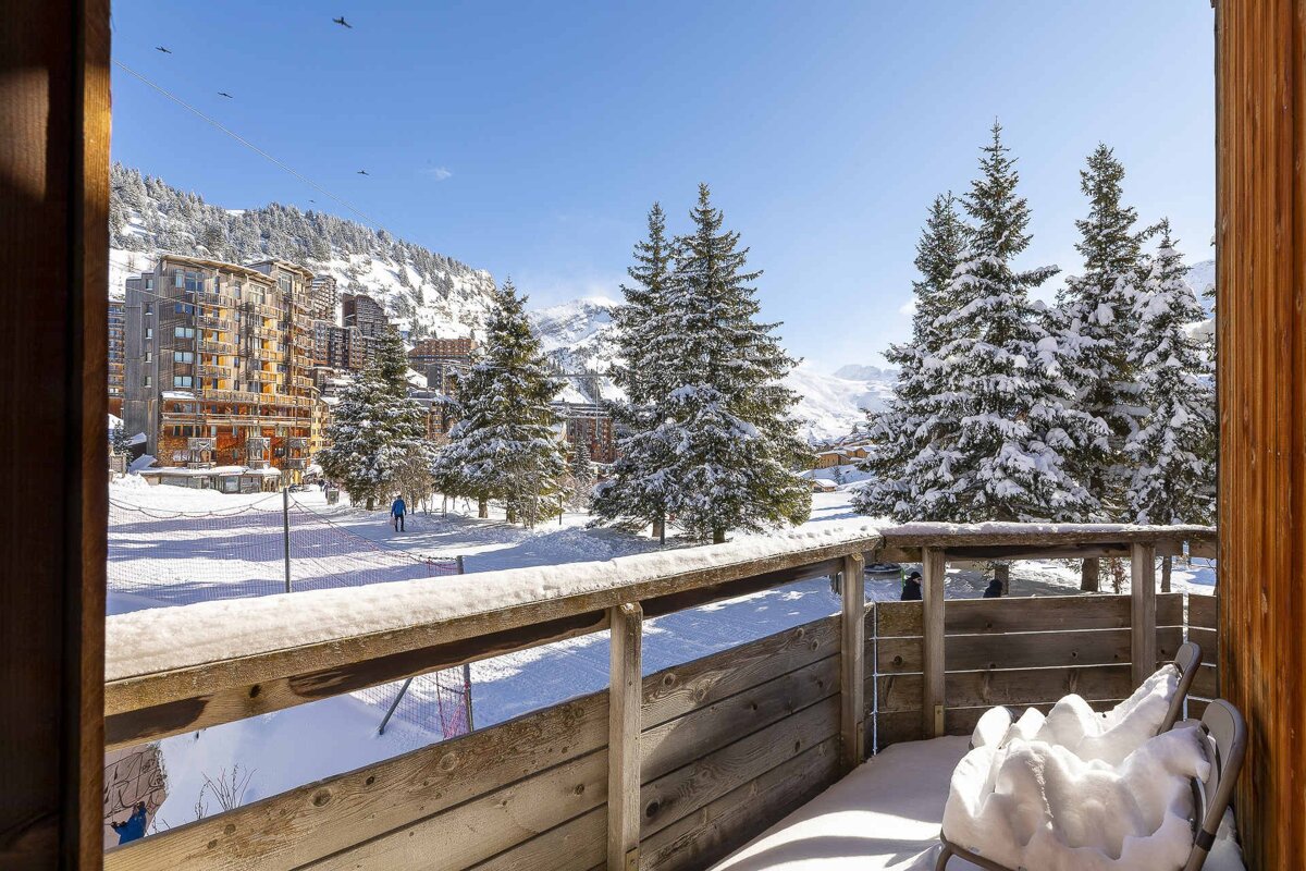 View from a snowy balcony of a winter mountain resort with snow-covered pine trees, apartment buildings, and distant peaks under a clear blue sky.