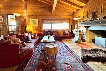 A cozy, rustic cabin living room with wooden walls, red sofas, a large patterned rug, warm lighting, and a fireplace.