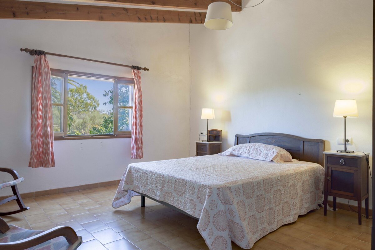 Bright bedroom with a bed, nightstands, window with tree view, tiled floor, wood beams, and a partial rocking chair.