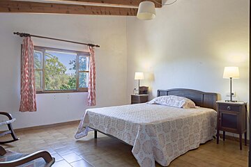 Bright bedroom with a bed, nightstands, window with tree view, tiled floor, wood beams, and a partial rocking chair.