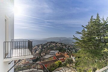 Sunny balcony view of a hillside town with distant mountains, trees, and a bright blue sky.