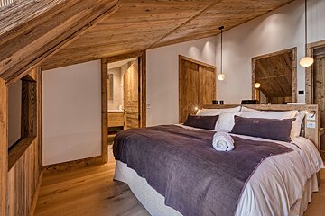 A cozy, rustic bedroom featuring natural wood walls, sloped ceiling, and a large bed with a purple throw, leading to an en-suite bathroom.