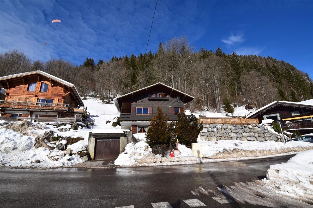 Snowy mountain village with wooden chalets, forested slopes, a wet road, and two paragliders flying high in the blue sky.