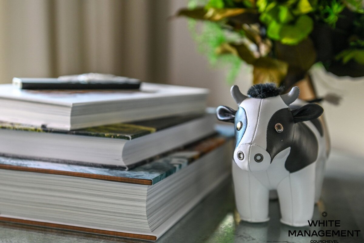 A black and white cow figurine sits next to a stack of books, with a blurred green plant in the background.