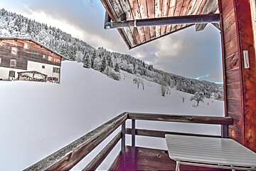 View from a rustic wooden balcony showcasing a snow-covered mountain with evergreen trees, a distant traditional house, and cloudy skies.