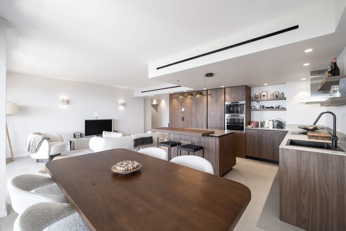 Modern open-plan apartment featuring a wooden dining table, sleek kitchen with dark cabinets, and a bright living area.
