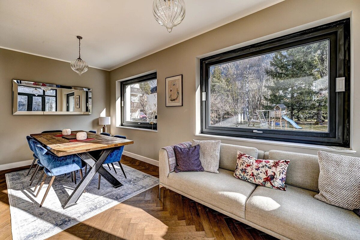 A bright, modern room with a dining area (wood table, blue chairs) and living space (sofa). Large windows offer views of a garden with a playground.