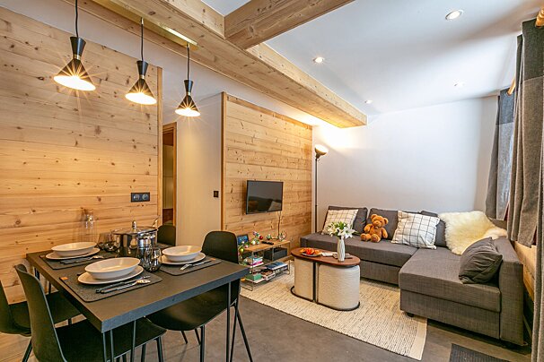 A modern open-plan living/dining space with wood-paneled walls, exposed beams, a grey sectional sofa, black dining table, and contemporary lighting.
