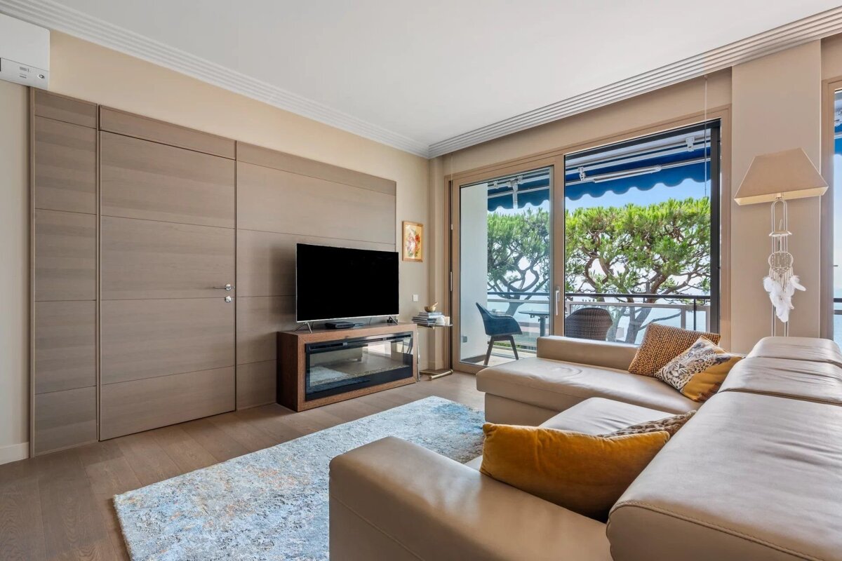 A modern living room featuring a beige leather sofa, a TV stand, wood paneled walls, and sliding glass doors leading to a balcony.