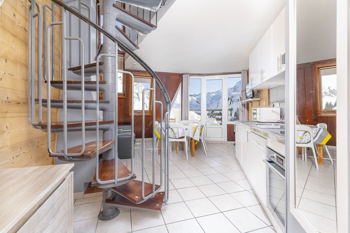 A chalet interior with a spiral staircase, compact kitchen/dining, and large windows offering a breathtaking snowy mountain view.