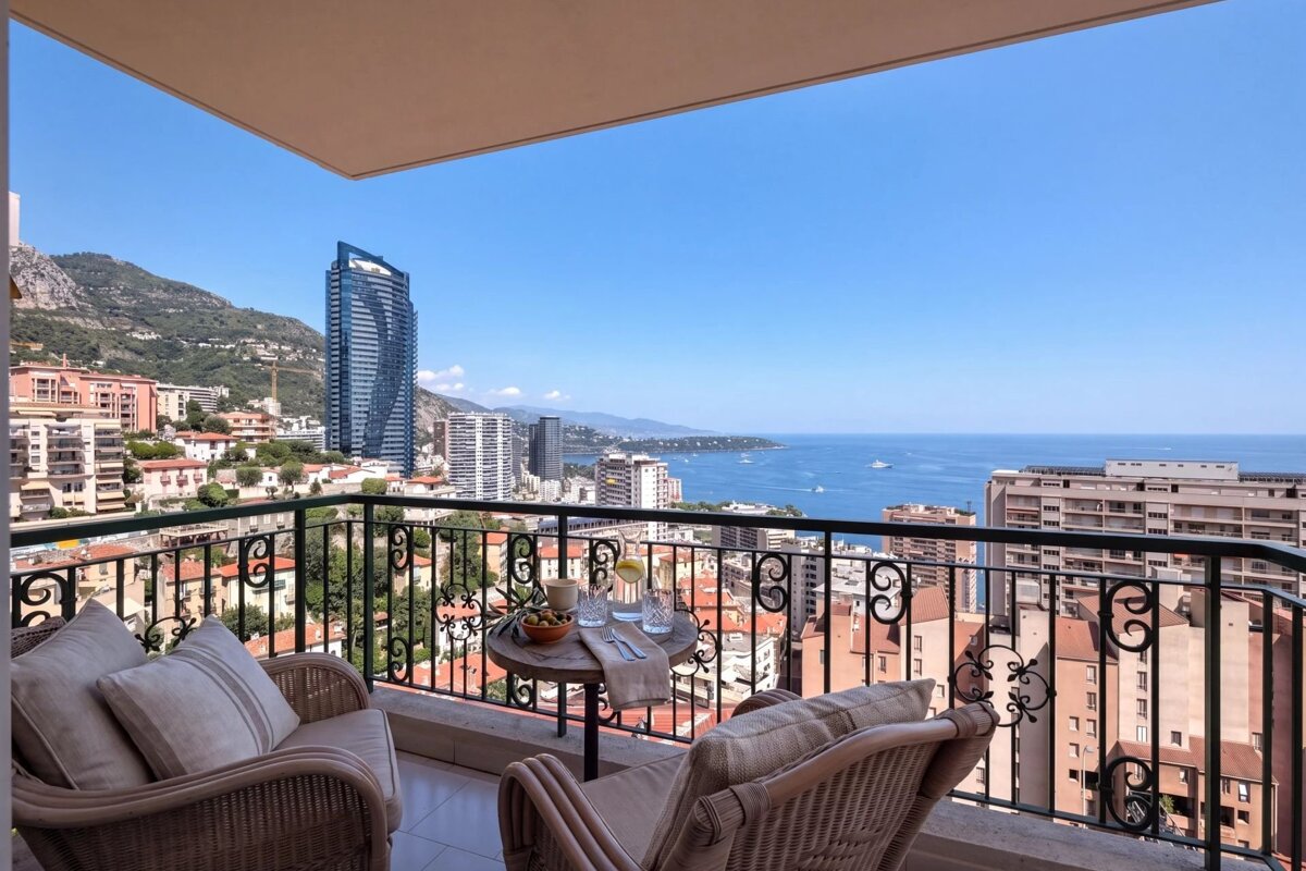 A balcony with comfortable chairs overlooking the city of Monaco and the Mediterranean Sea under a bright blue sky.