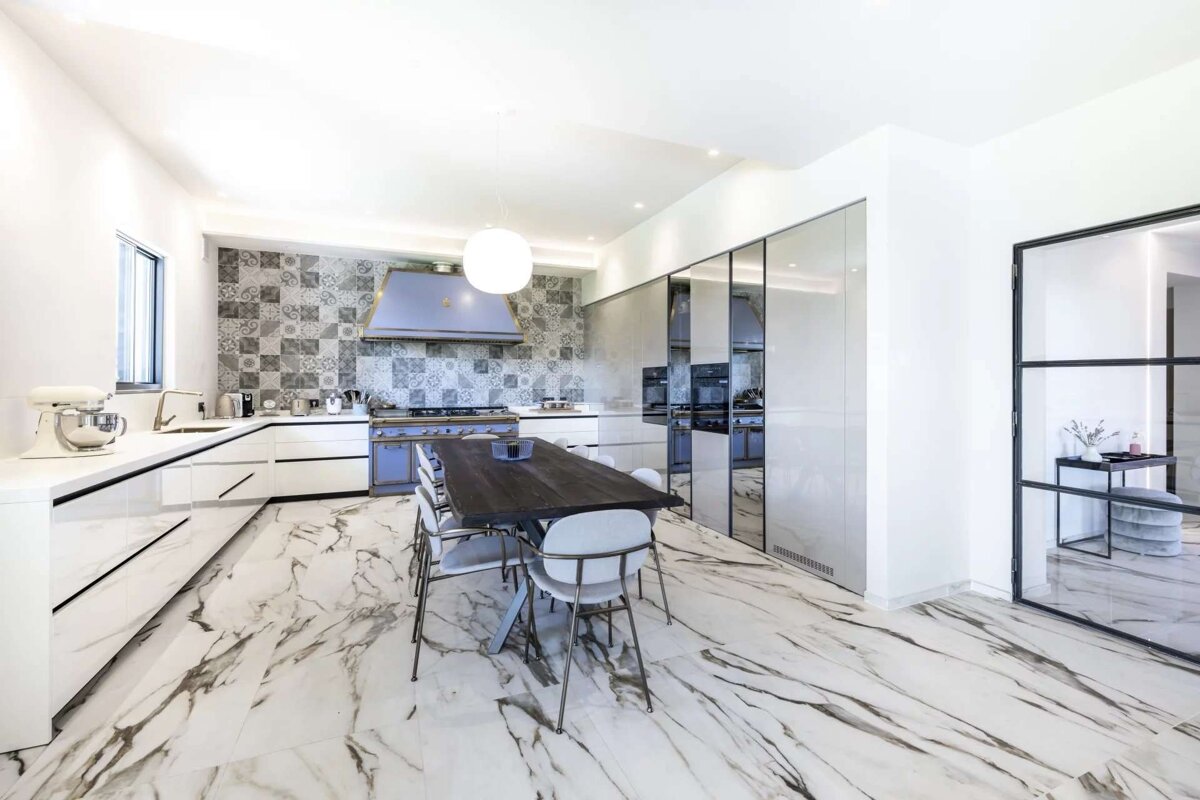 A bright, modern kitchen with white cabinets, marble-patterned floor, a tiled backsplash, and a striking lilac oven. A long dining table sits centrally.