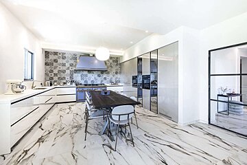 A bright, modern kitchen with white cabinets, marble-patterned floor, a tiled backsplash, and a striking lilac oven. A long dining table sits centrally.