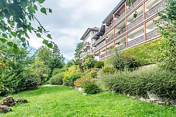 A multi-story building with balconies sits above a lush, terraced garden and green lawn under a cloudy sky.