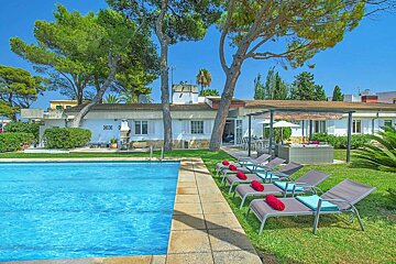 A sunny villa with a clear blue swimming pool, several sun loungers on a green lawn, and tall trees under a bright sky.