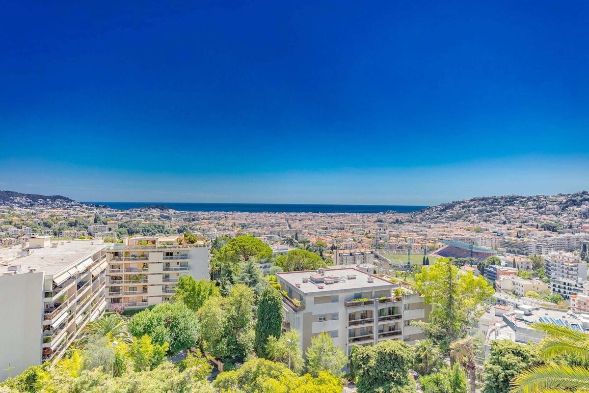 A high-angle view of a sunny coastal city with numerous buildings, green trees, and distant ocean under a vast, clear blue sky.