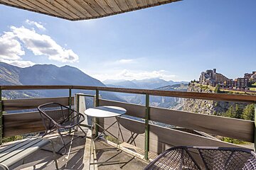 Sunny balcony with chairs and a table overlooking a vast mountain valley and cliffside resort under a blue sky.