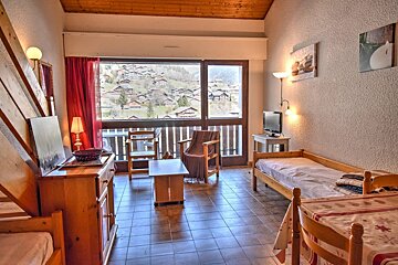 A rustic room with wood furniture, tiled floor, and two beds, featuring a large window with a scenic view of a hillside village.