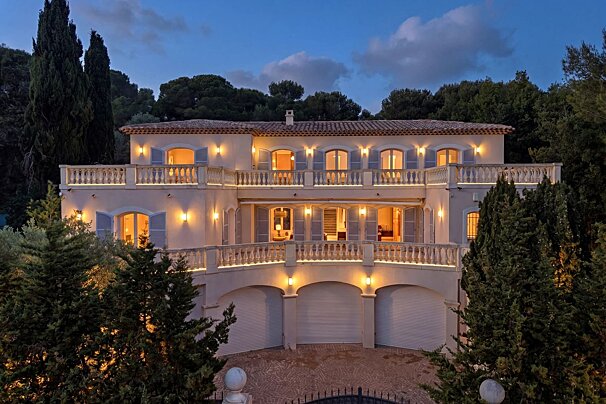 An elegant multi-story villa with balconies, arched windows, and soft lights, nestled in lush trees at dusk under a twilight sky.
