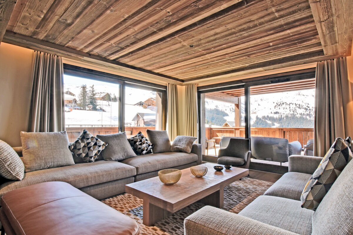 A luxurious ski chalet living room with wooden ceilings, comfy sofas, and large windows revealing a scenic snowy mountain landscape.