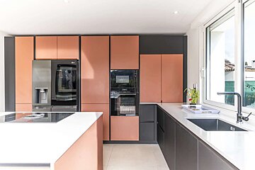Modern kitchen with rose gold and dark grey cabinets, white countertops, built-in appliances, and a central island. Bright window over the sink.