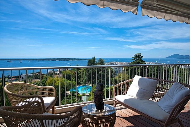 Sunny balcony overlooking a beautiful blue sea, with wicker furniture, a pool below, and a clear sky.