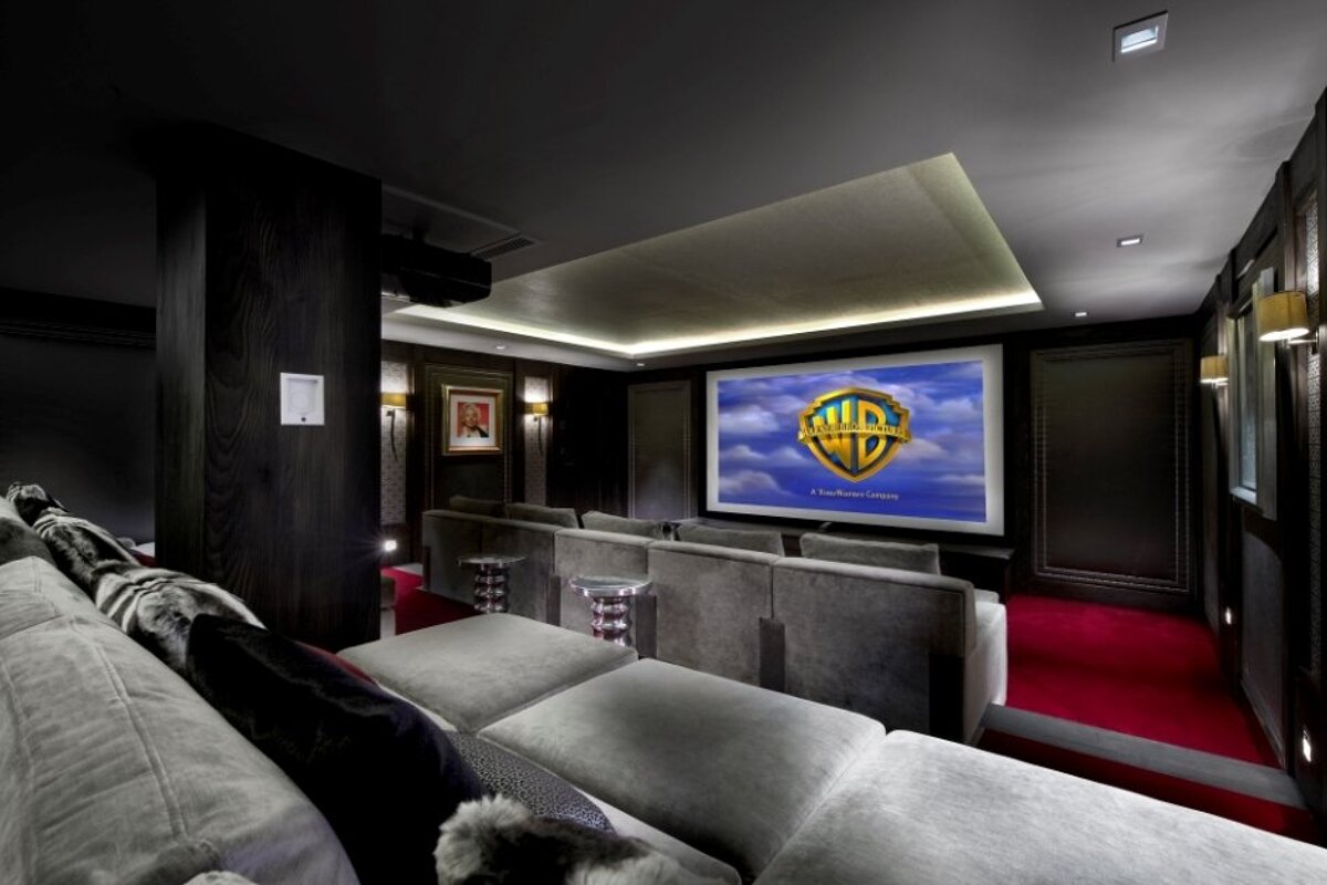 Luxurious home theater with a large screen showing the Warner Bros. logo, dark wood walls, red carpet, and comfortable gray seating.