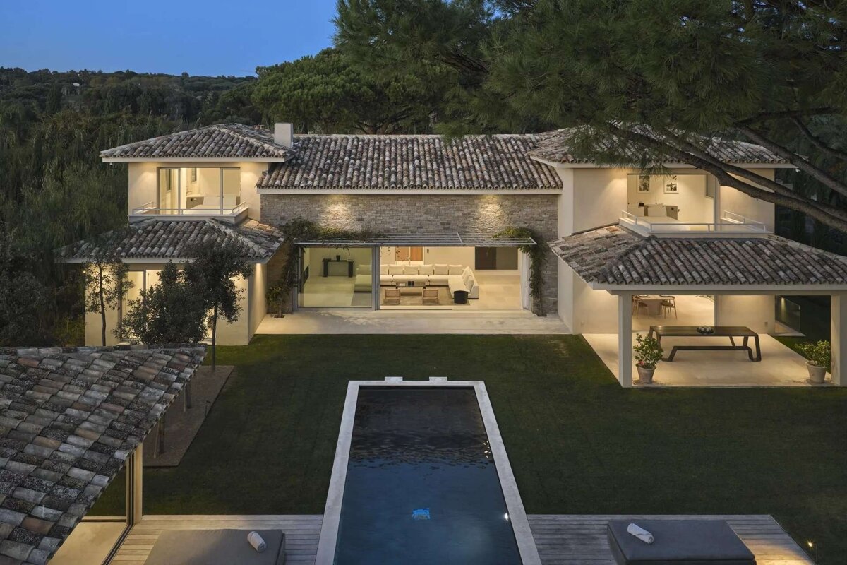 Luxurious villa at dusk with illuminated interiors, a dark pool, and a green lawn, surrounded by lush trees.