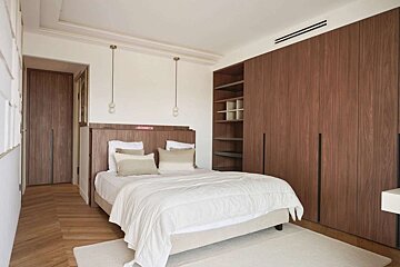 Modern bedroom with white walls, large wooden wardrobes, a light-colored bed with white bedding, herringbone floor, and two hanging pendant lights.