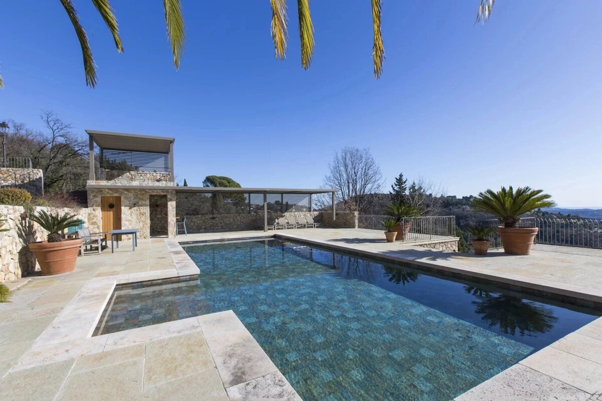 A sunny outdoor swimming pool with a stone patio, surrounded by potted plants and a clear blue sky.