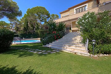 A large villa with grand outdoor stairs leading to a patio, vibrant green lawn, and a sparkling blue swimming pool, all surrounded by lush trees under a sunny sky.
