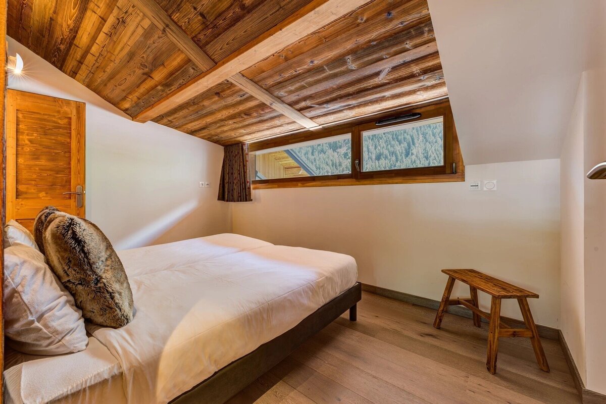 Cozy cabin bedroom with a sloped wooden ceiling, a white bed, a rustic bench, and a window looking out at a pine forest.