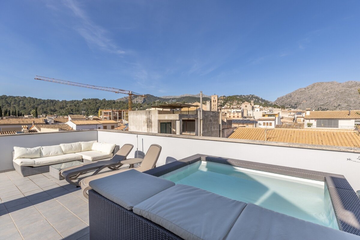 Sunny rooftop terrace with a plunge pool, lounge seating, and panoramic views of a town, mountains, and clear blue sky.