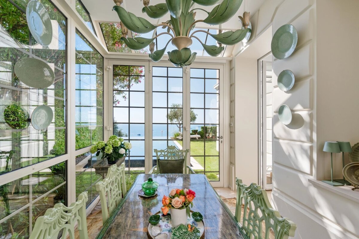 Bright sunroom dining area featuring a green leaf chandelier, carved chairs, and glass walls overlooking a garden and ocean.