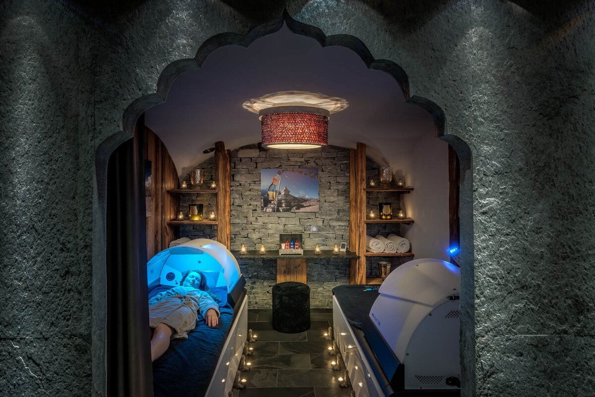 Cozy spa room viewed through a stone archway, featuring a woman relaxing in a blue-lit therapy pod surrounded by warm candlelight.