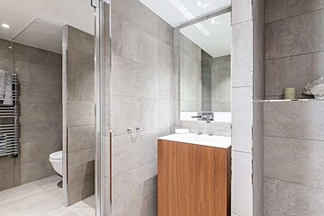 A modern bathroom with grey tiled walls, wooden vanity, mirror, and a partial view of a toilet and heated towel rail behind a shower screen.