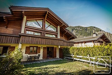 A grand wooden chalet with large windows overlooks a green lawn and patio, set against a majestic mountain range under a clear blue sky.